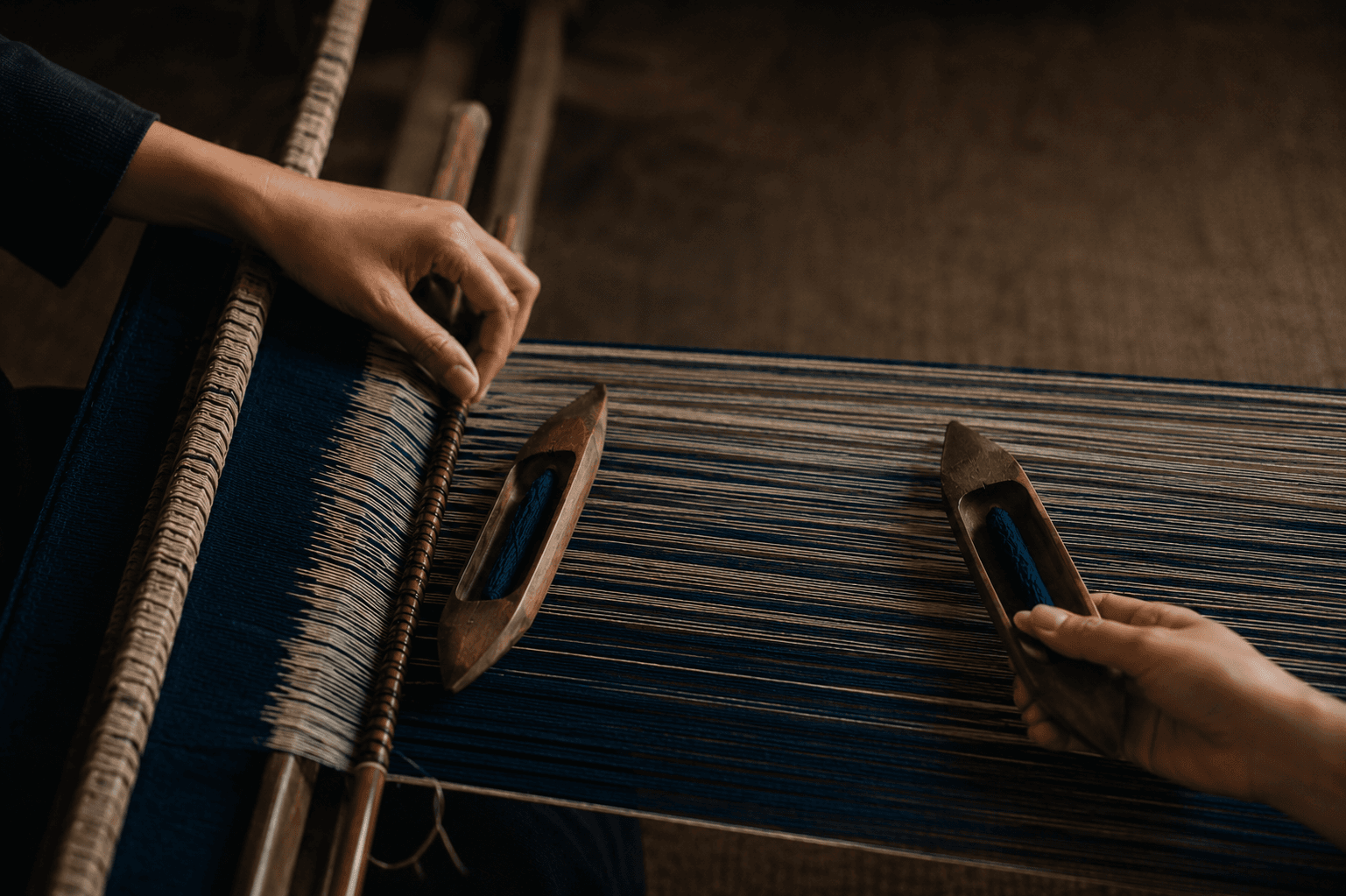 Two hands passing a shuttle of indigo thread across a backstrap loom.